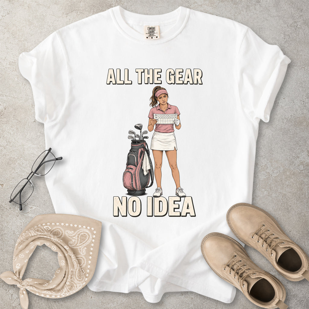 White t-shirt with a graphic of a golfer and text 'All the gear no idea' on a gray surface, surrounded by glasses, bandana, and shoes.