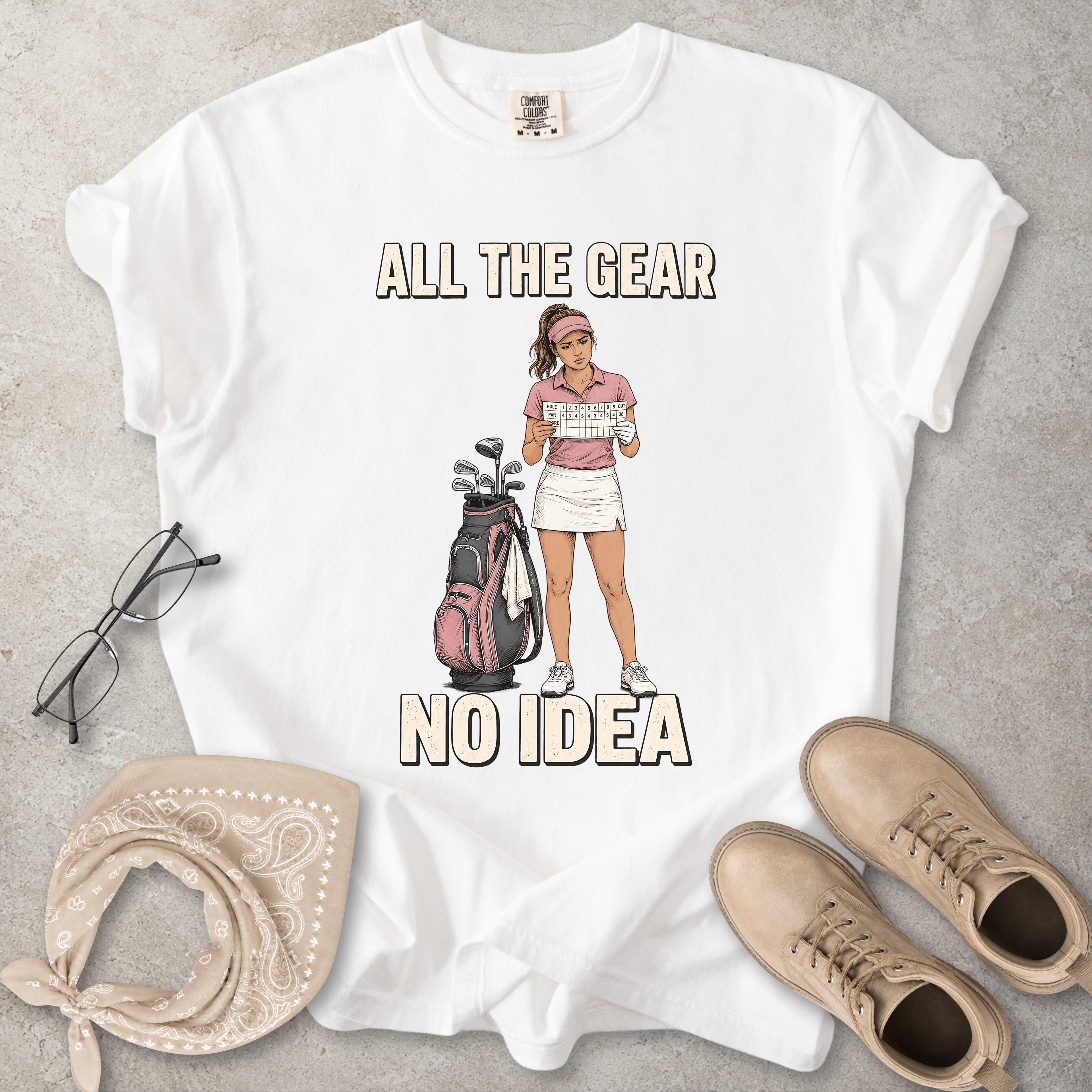 White t-shirt with a graphic of a golfer and text 'All the gear no idea' on a gray surface, surrounded by glasses, bandana, and shoes.