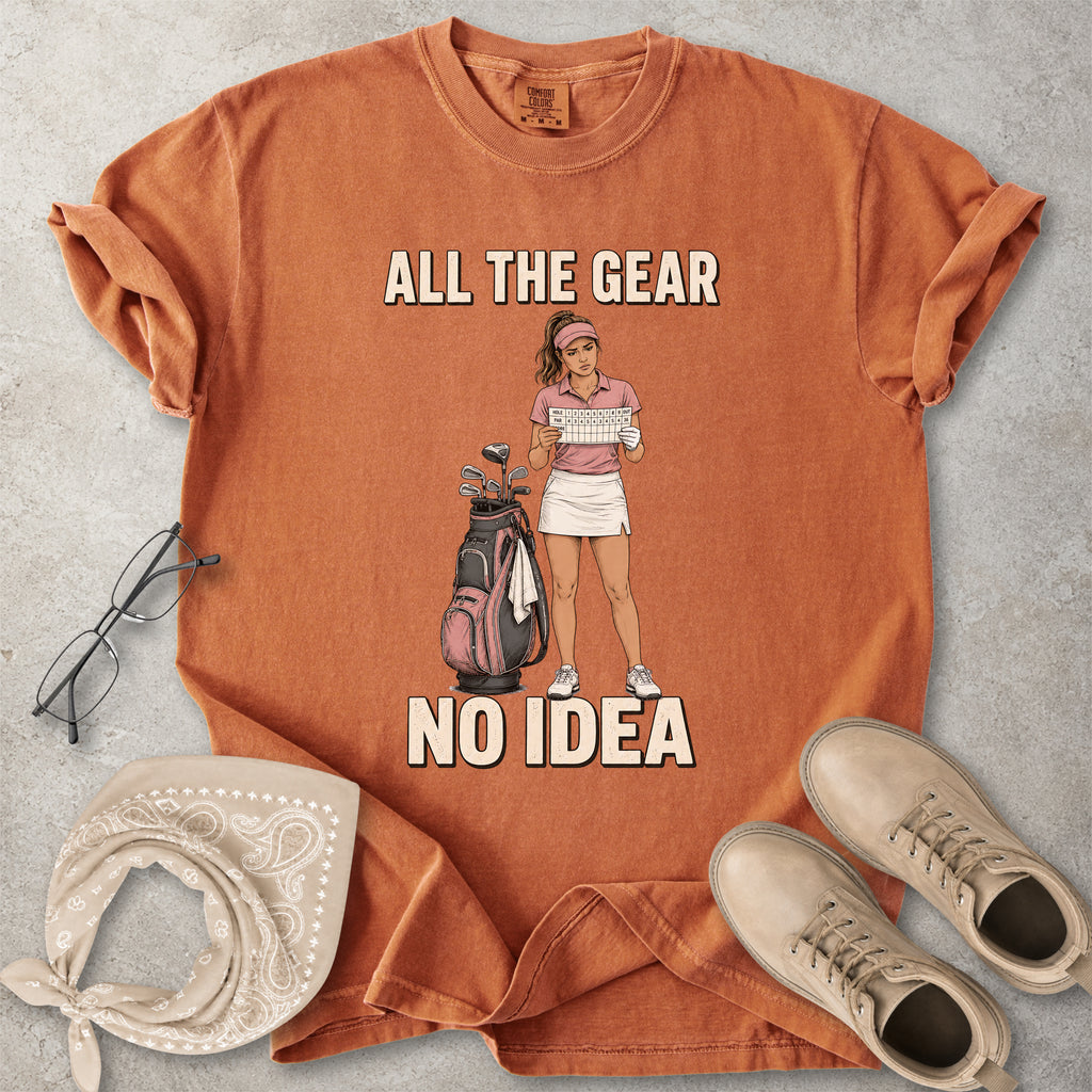 Yam Brown t-shirt with graphic design of a woman holding a golf club and text 'All the gear no idea' on a gray surface with accessories.