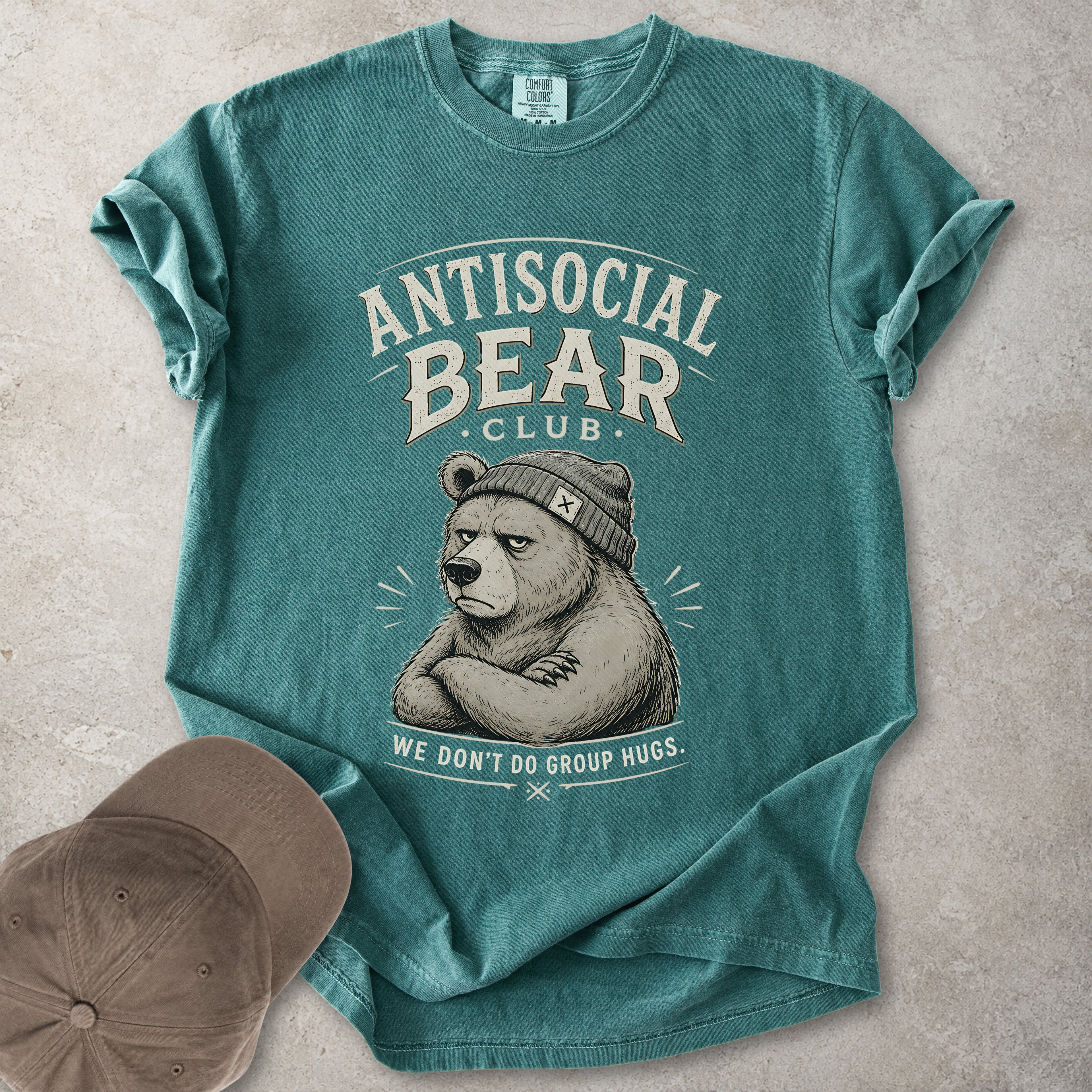 Green Comfort Colors graphic T-shirt with Antisocial Bear Club design showing a grumpy bear in a beanie and text reading we don’t do group hugs, styled with cap and tan boots