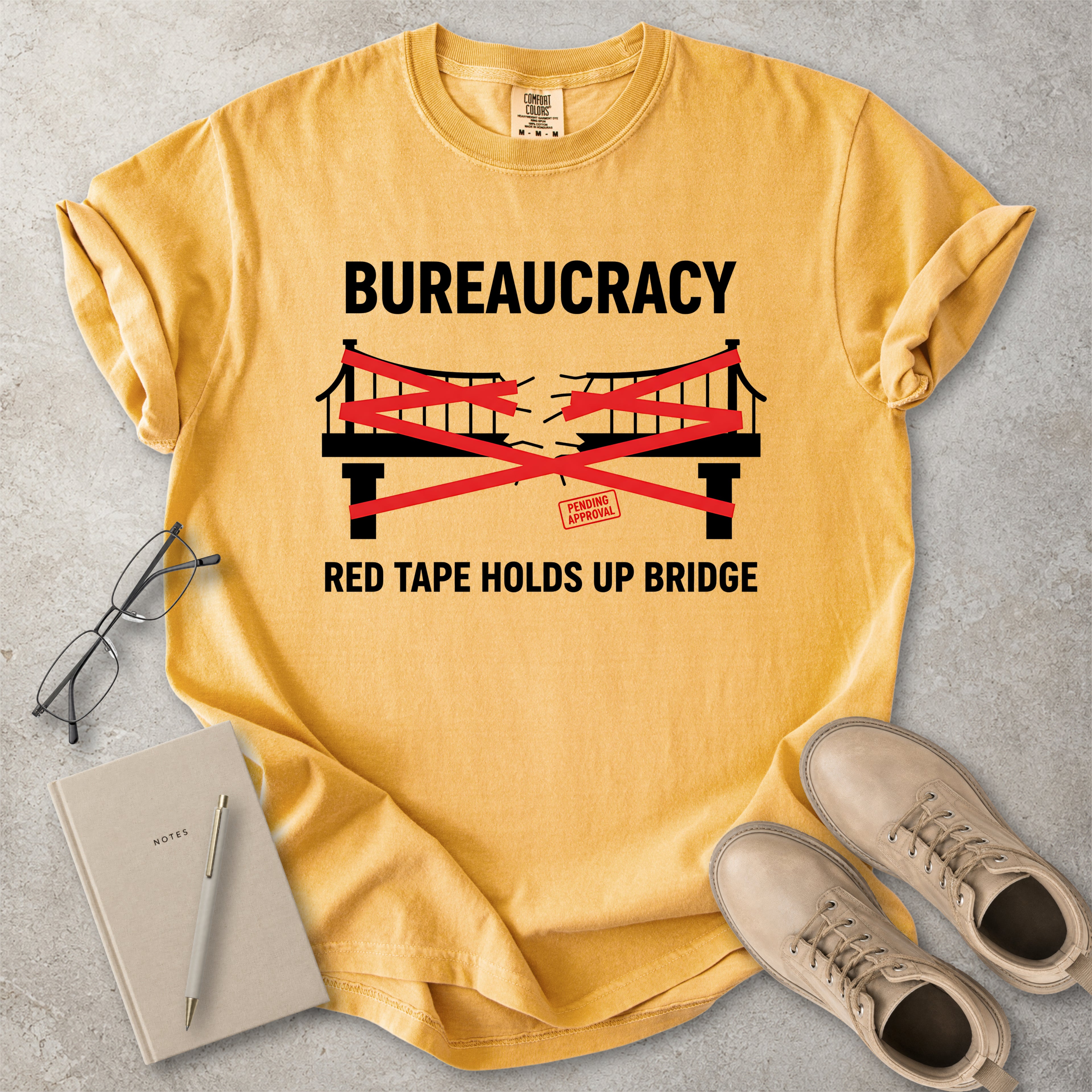 Mustard yellow Comfort Colors graphic T-shirt with bureaucracy red tape holds up bridge design, styled with notebook, glasses and tan boots