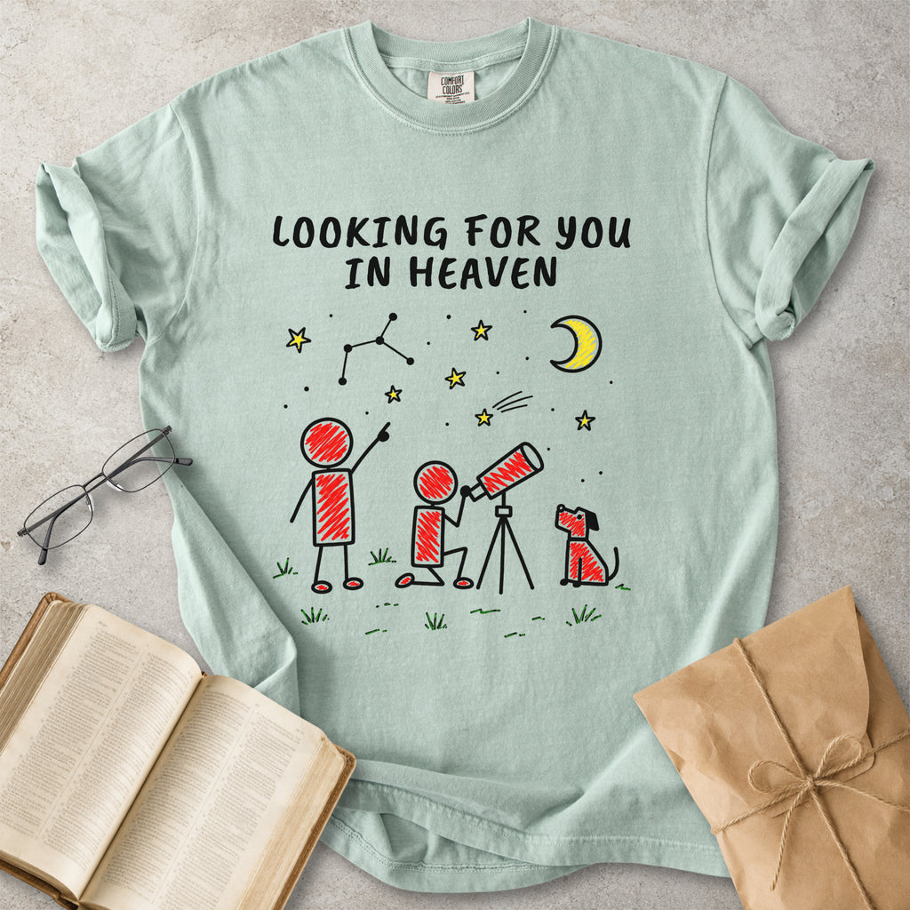A mint green graphic t-shirt laid flat on a textured gray surface, surrounded by lifestyle props including an open book, eyeglasses, and a brown paper gift bag. The t-shirt features a cartoon graphic of two children and a dog looking through a telescope under a crescent moon and stars, with the text 'LOOKING FOR YOU IN HEAVEN'.