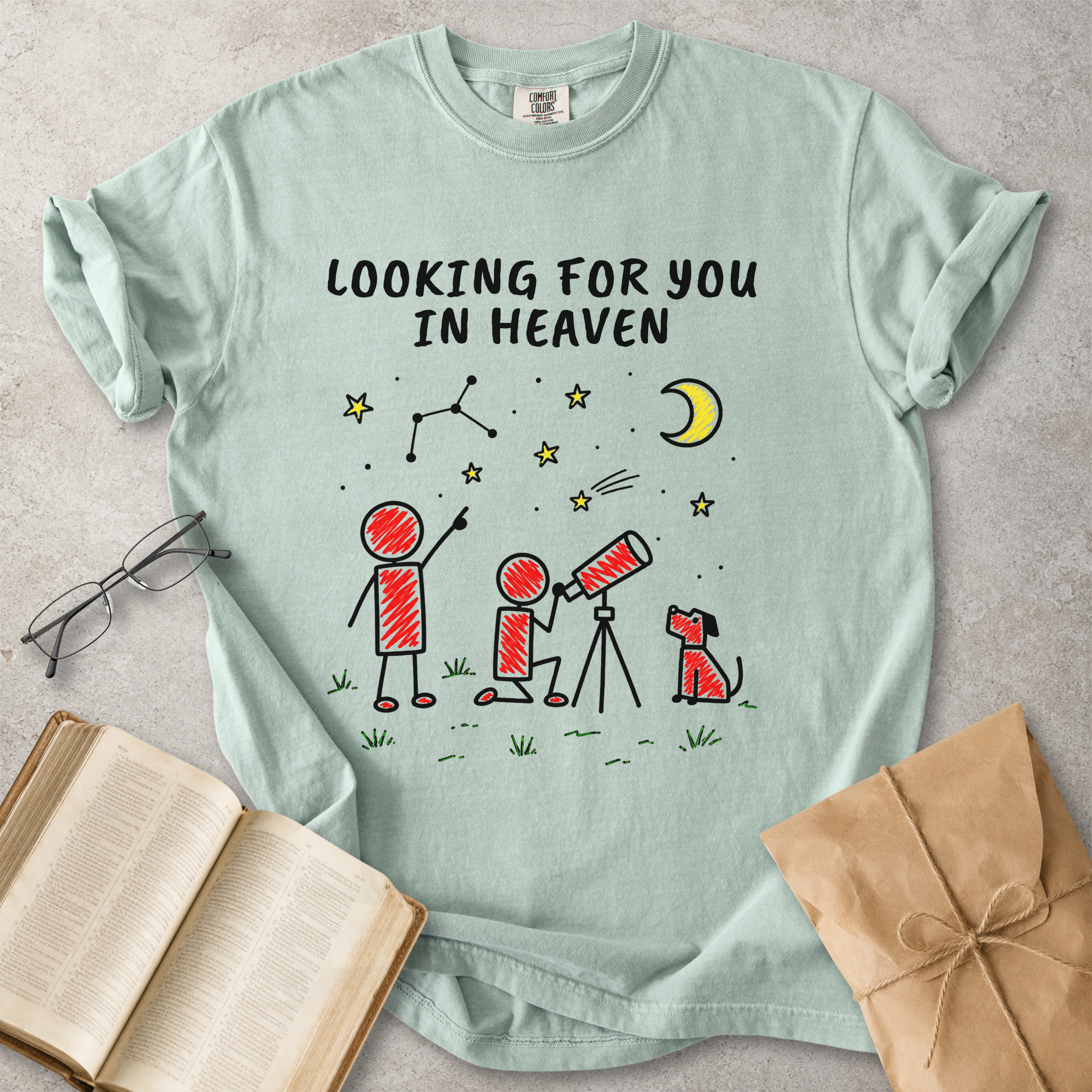 A mint green graphic t-shirt laid flat on a textured gray surface, surrounded by lifestyle props including an open book, eyeglasses, and a brown paper gift bag. The t-shirt features a cartoon graphic of two children and a dog looking through a telescope under a crescent moon and stars, with the text 'LOOKING FOR YOU IN HEAVEN'.
