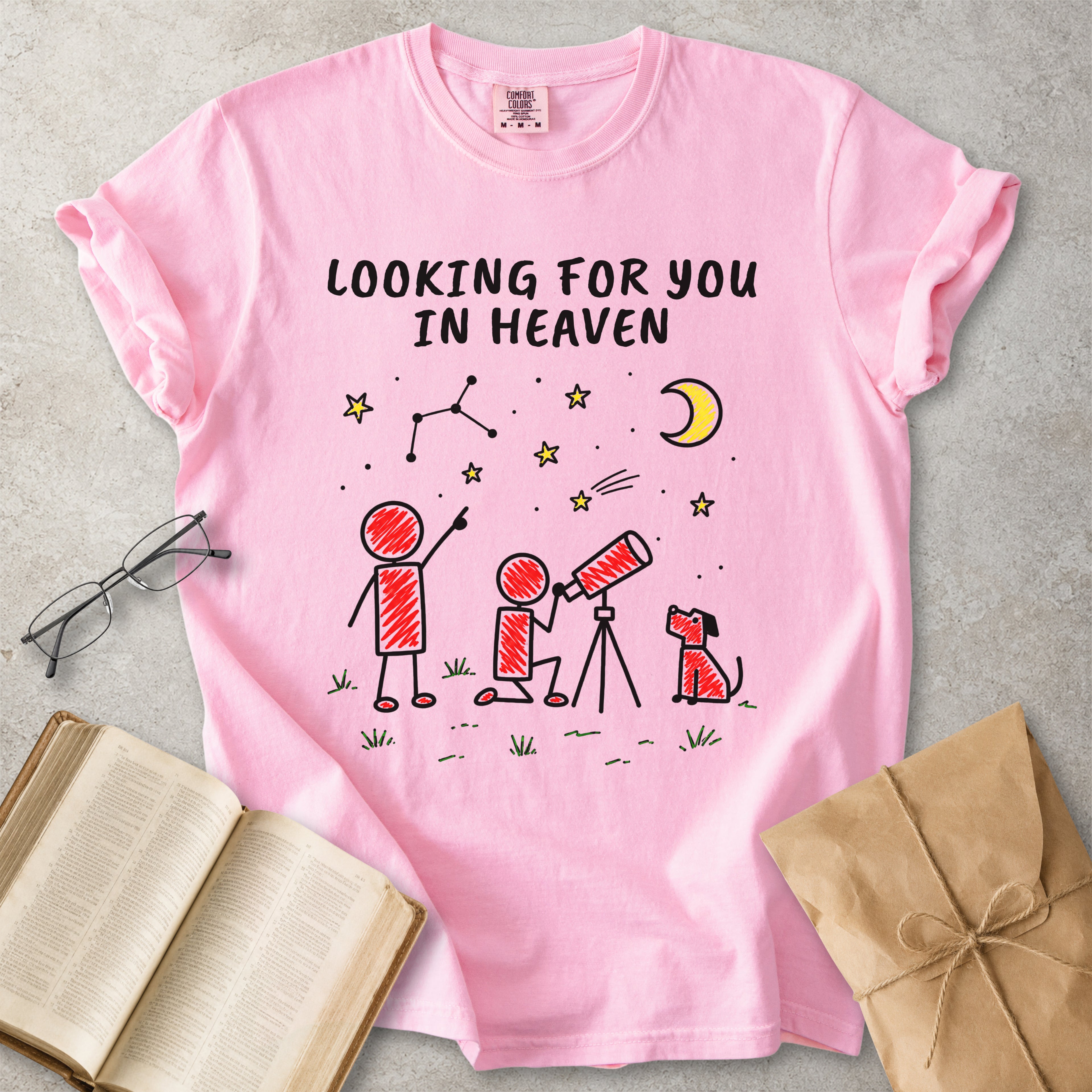 A pink short-sleeve graphic T-shirt laid flat on a light gray surface, surrounded by styled props including an open book, eyeglasses, and a brown paper envelope. The shirt has black text reading 'LOOKING FOR YOU IN HEAVEN' above a simple red, black, and yellow sketch of two people looking up at a crescent moon and stars.