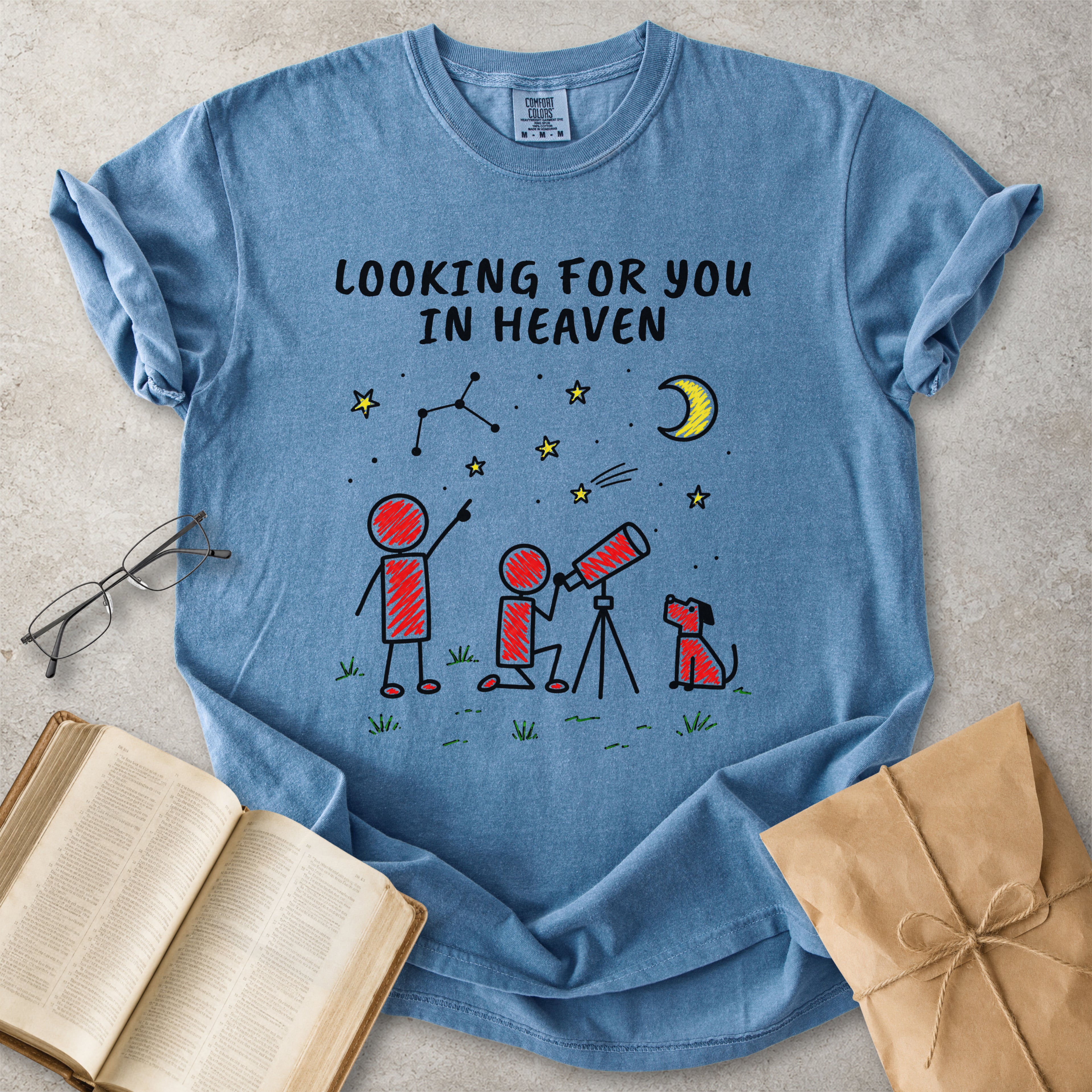 A light blue graphic t-shirt laid flat on a textured gray surface, surrounded by styled props including an open book, eyeglasses, and a brown paper gift bag. The t-shirt features a graphic of stick figures looking up at a crescent moon and stars with the text 'LOOKING FOR YOU IN HEAVEN'.
