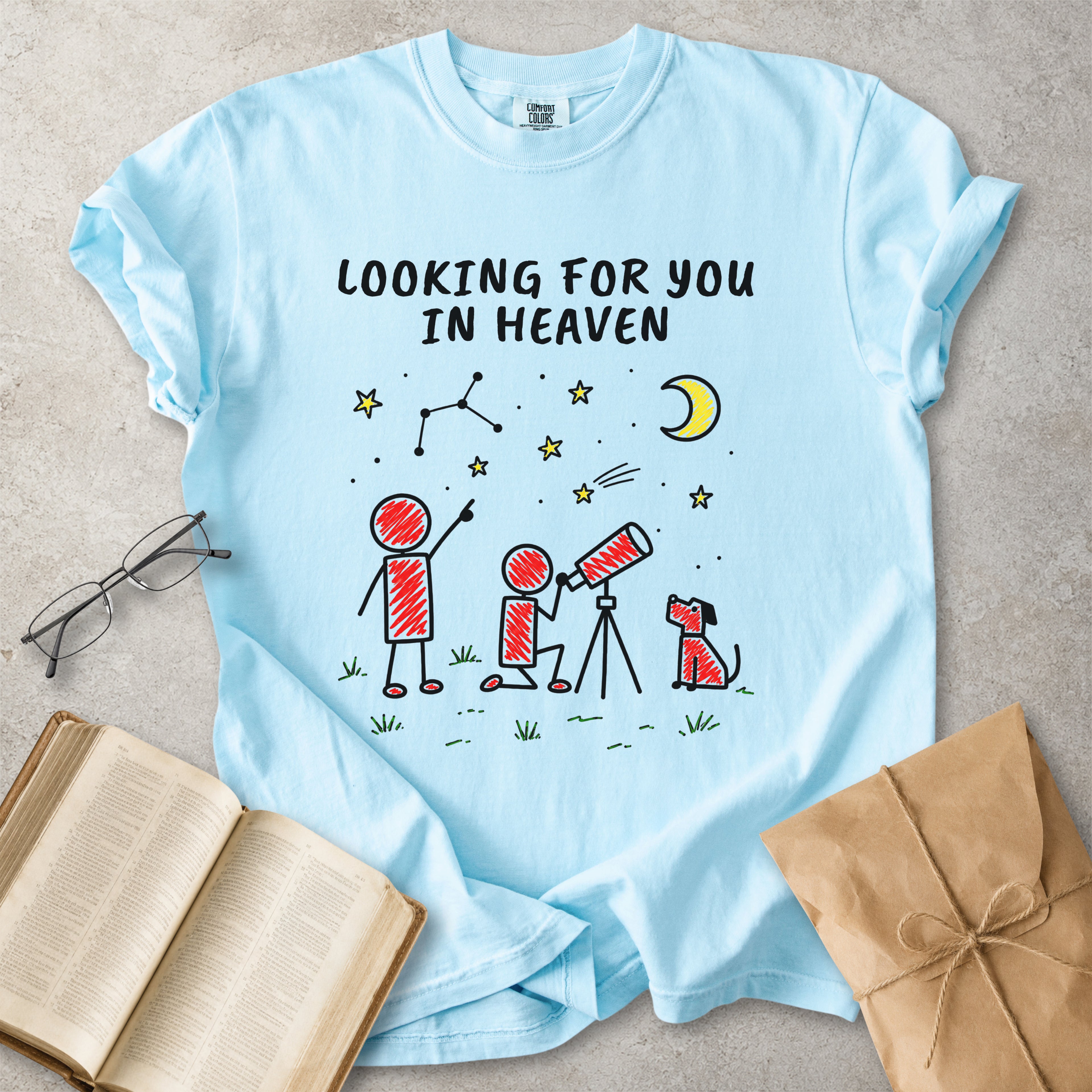 A light blue graphic t-shirt laid flat on a textured gray surface, surrounded by props including an open book, eyeglasses, and a brown paper gift bag. The t-shirt features a black hand-drawn style graphic of stick figures looking up at a crescent moon and stars, with the text 'LOOKING FOR YOU IN HEAVEN'.