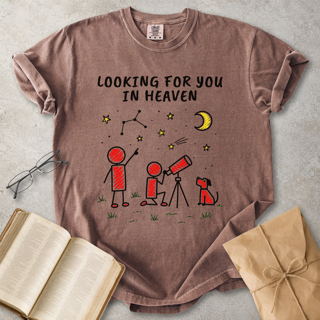 A brown short-sleeve graphic T-shirt laid flat on a light gray surface, surrounded by an open book, eyeglasses, and a kraft paper gift bag. The shirt has a printed design with the text 'LOOKING FOR YOU IN HEAVEN' above stick-figure children, stars, a crescent moon, and a small dog.