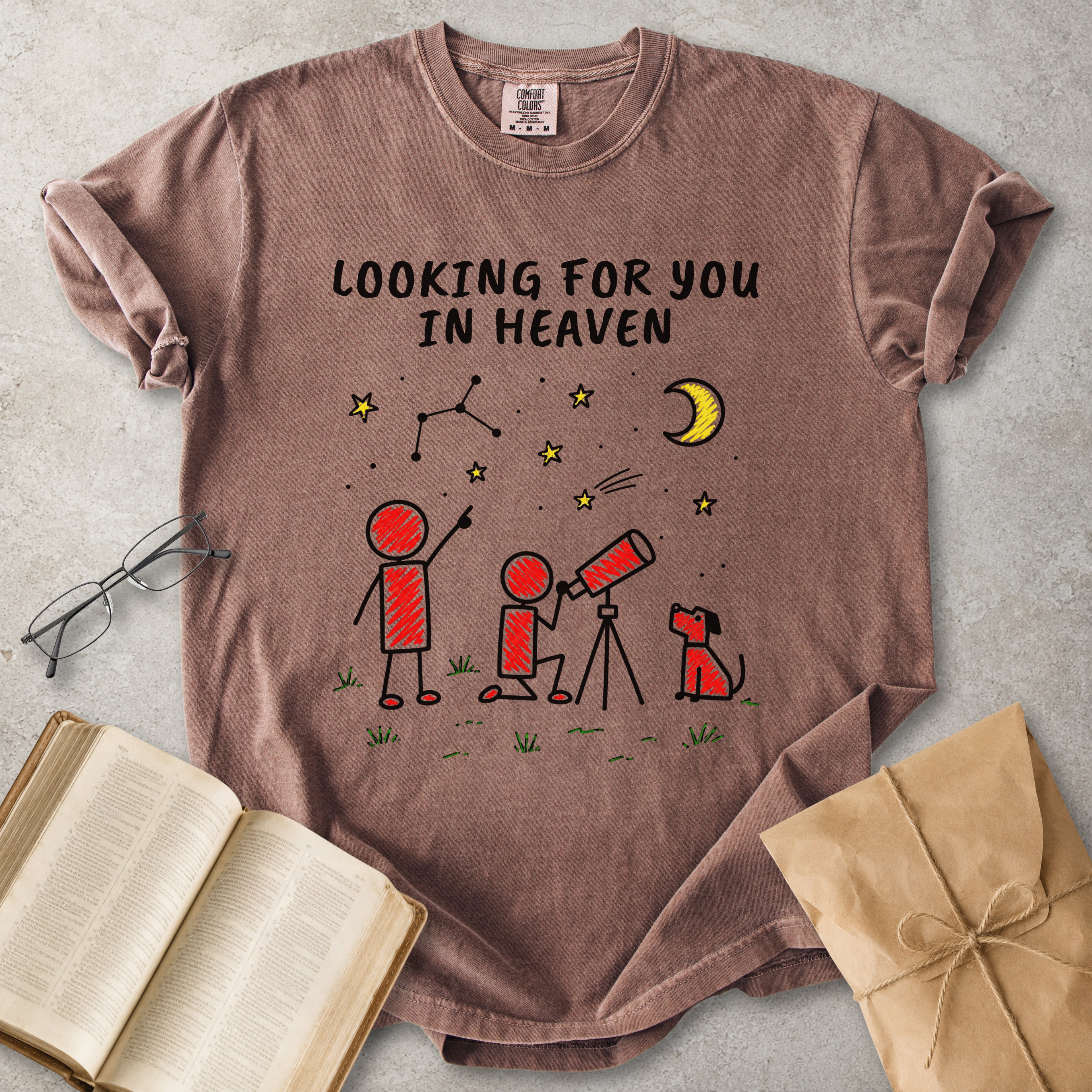 A brown short-sleeve graphic T-shirt laid flat on a light gray surface, surrounded by an open book, eyeglasses, and a kraft paper gift bag. The shirt has a printed design with the text 'LOOKING FOR YOU IN HEAVEN' above stick-figure children, stars, a crescent moon, and a small dog.
