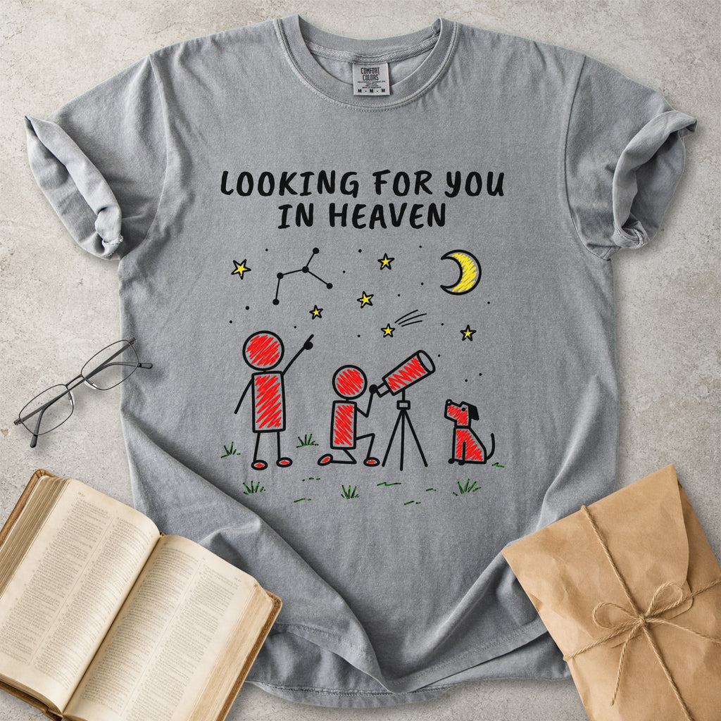 A gray short-sleeve t-shirt with a graphic print of stick figures looking up at a crescent moon and stars, with the text 'LOOKING FOR YOU IN HEAVEN'. The shirt is styled in a flat-lay arrangement on a textured gray surface with an open book, eyeglasses, and a brown paper gift bag.