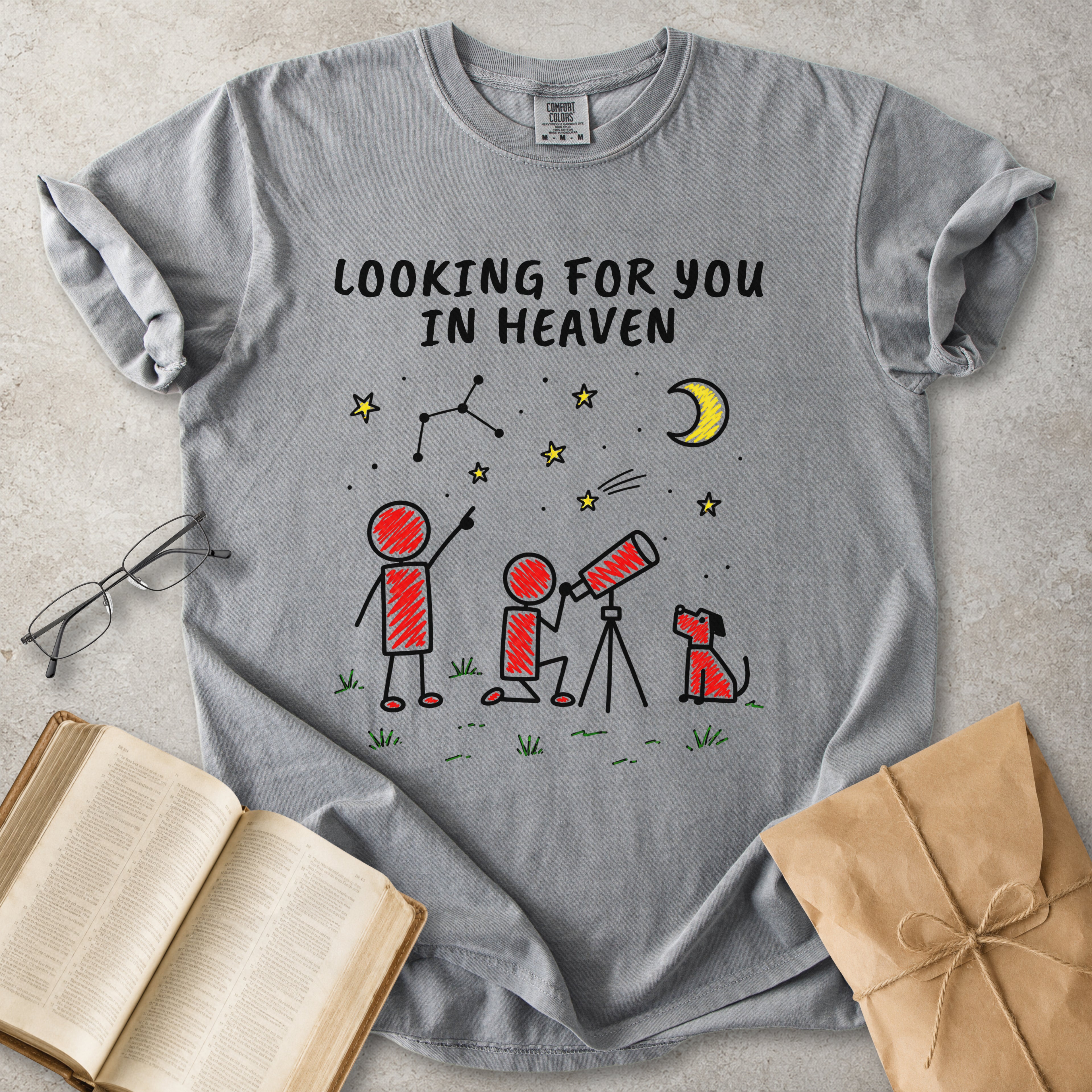 A gray short-sleeve t-shirt with a graphic print of stick figures looking up at a crescent moon and stars, with the text 'LOOKING FOR YOU IN HEAVEN'. The shirt is styled in a flat-lay arrangement on a textured gray surface with an open book, eyeglasses, and a brown paper gift bag.