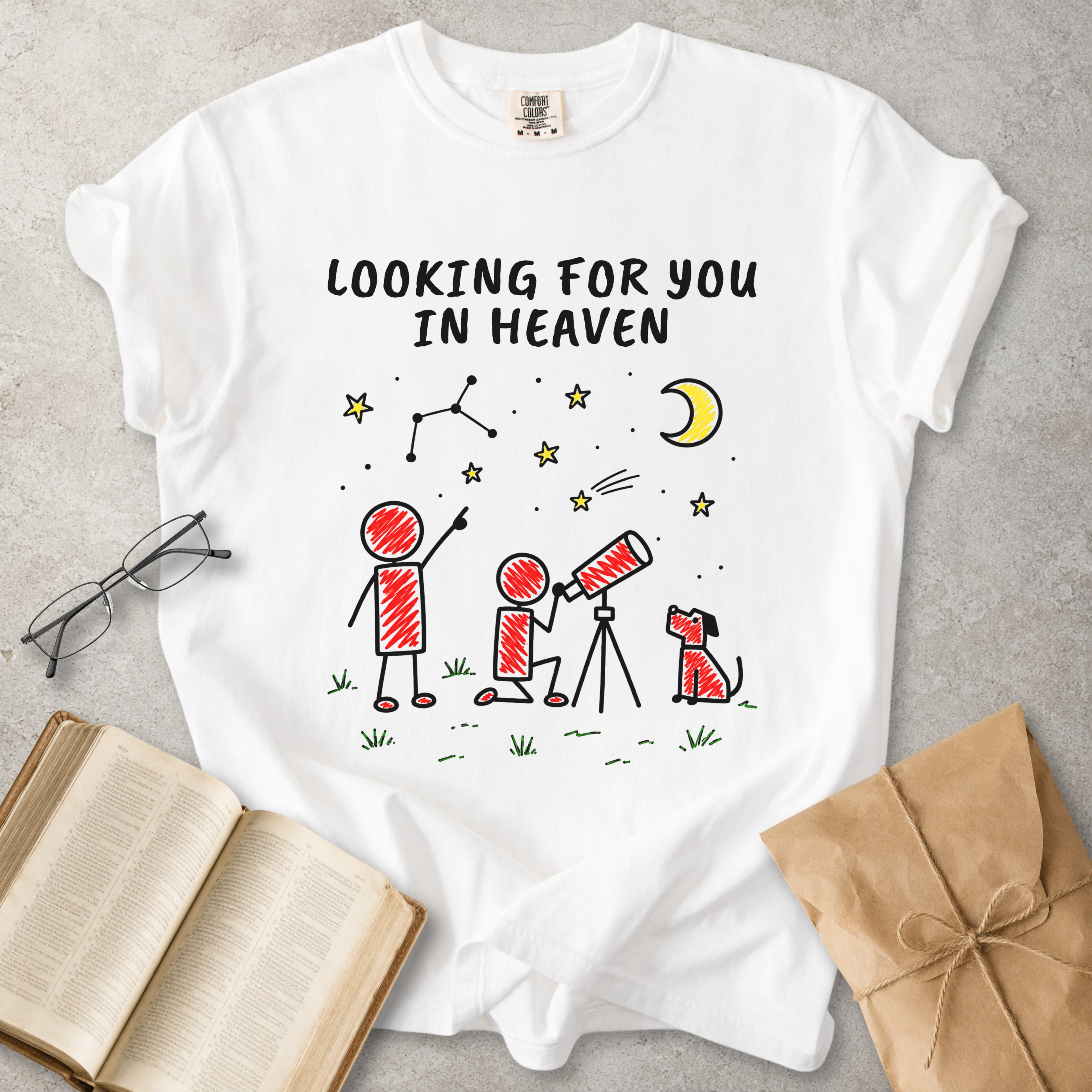 A white graphic t-shirt laid flat on a gray textured surface, surrounded by styled props including an open book, eyeglasses, and a brown paper gift bag. The t-shirt features a hand-drawn style illustration of stick figures, a dog, stars, a crescent moon, and constellations with the text 'LOOKING FOR YOU IN HEAVEN' printed in black.