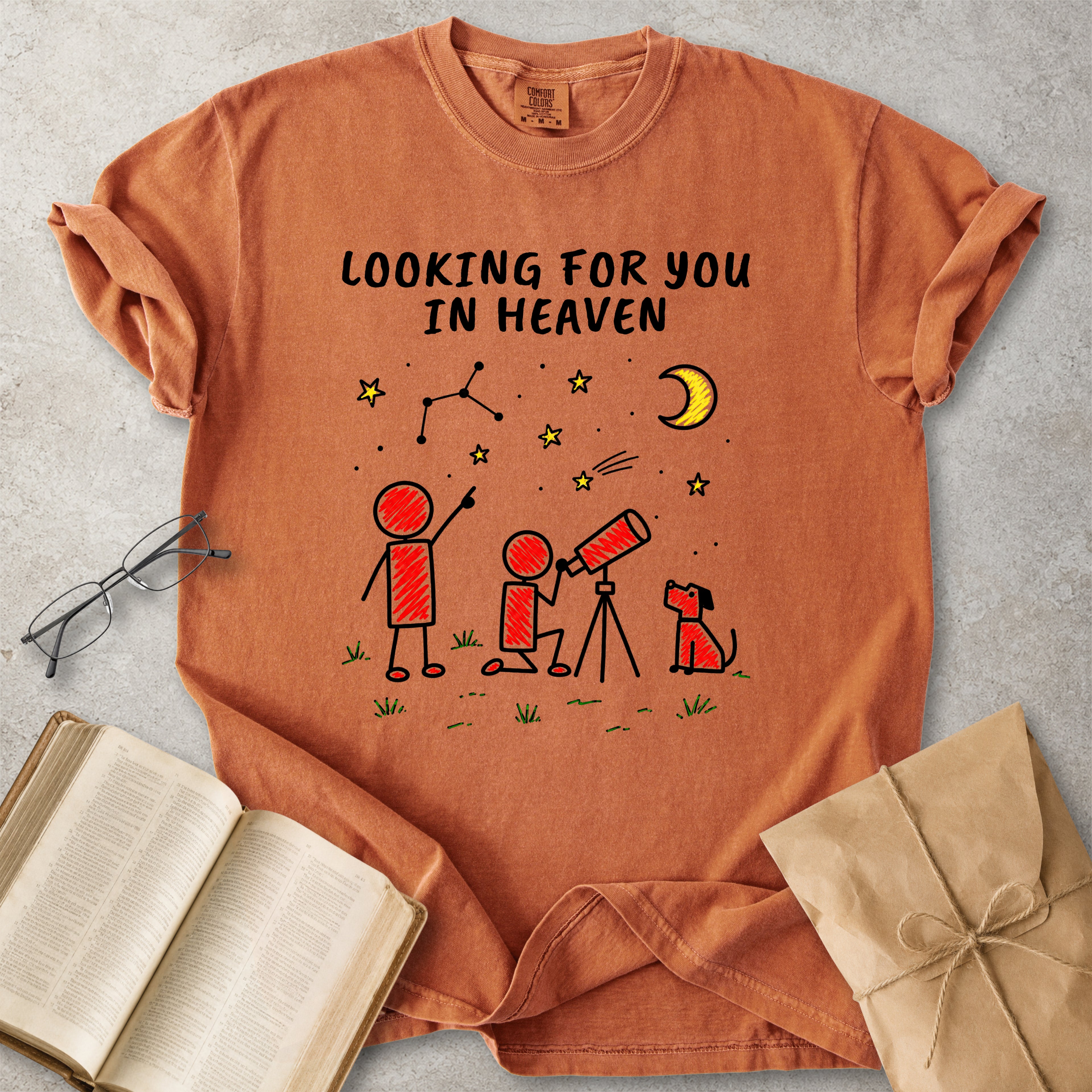 An orange short-sleeve graphic T-shirt laid flat on a light gray textured surface, surrounded by styled props including an open book, eyeglasses, and a wrapped package. The shirt has a printed celestial scene with the text 'LOOKING FOR YOU IN HEAVEN' above stick figures, stars, a crescent moon, and a constellation.
